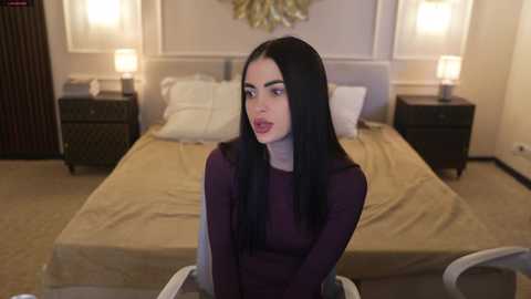 Video of a woman with long black hair and fair skin, wearing a long-sleeved dark purple dress, sitting on a white chair in a modern bedroom with beige walls, a bed, and two nightstands with lamps.