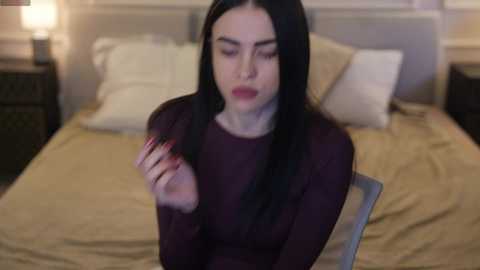 Media: A video of a pale woman with long black hair, wearing a maroon sweater, sitting on a beige bed with white pillows. She's holding her hair and looks distressed. The background shows a nightstand with a lamp.