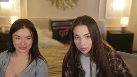 Media: Video of two young women with long dark hair, smiling, sitting side by side on a bed in a warmly lit, tidy bedroom.