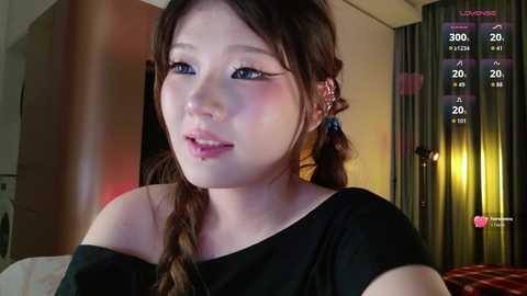 A video of an East Asian woman with fair skin and long brown hair styled in twin braids, wearing a black off-shoulder top, in a dimly lit room with a red curtain and a wooden door.