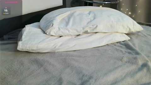 Video of a bed with two white pillows on a grey blanket, set against a dark wall with a small circular nightstand beside it.