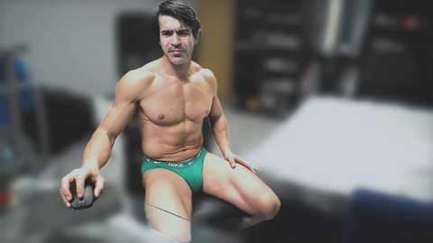 Video of a muscular, shirtless man with short dark hair, wearing green briefs, in a gym, holding a dumbbell, background blurred.