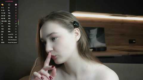 Video of a young, fair-skinned woman with straight, light brown hair, wearing a black hairpin, and holding a finger to her lips, indoors with a wooden wall and a mounted picture in the background.