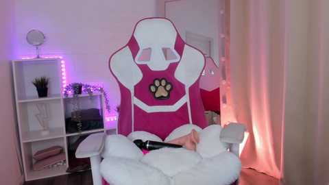 Video of a pink gaming chair with a paw print logo, white fluffy blanket, and beige curtains in a room with white shelves and purple LED strip lights.