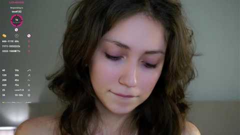 A video of a young woman with long, wavy brown hair, pale skin, and closed eyes, standing in a blurred indoor setting.