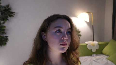 Video of a fair-skinned, brown-haired young woman with a contemplative expression, standing in a softly lit bedroom with a green sofa and a white fluffy pillow.