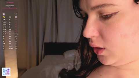 Video of a young woman with fair skin, dark hair, and dark eyeliner, looking introspective in a dimly lit bedroom.