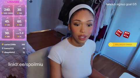 A young Black woman with a medium complexion, braids, and a white headband, wearing a white T-shirt, sits indoors with a wooden floor. On the left, a digital screen displays stats and emojis.