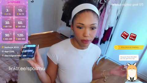 A video of a young Black woman with a light complexion, wearing a white headband and a tight white top, playing a mobile game on a wooden table in a cluttered room with hanging clothes.