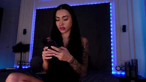 Video of a tattooed, dark-haired woman with long hair, wearing a black top, sitting on a bed, looking at a smartphone under blue LED lighting.