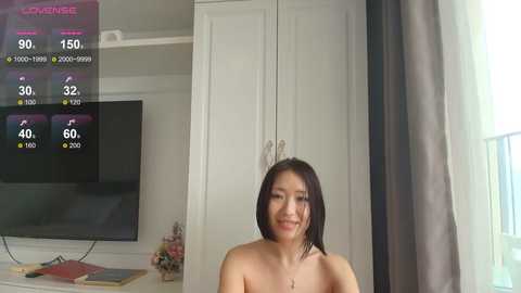 A video of an Asian woman with shoulder-length black hair, smiling, topless, standing in front of a white cabinet with a TV screen displaying scores.