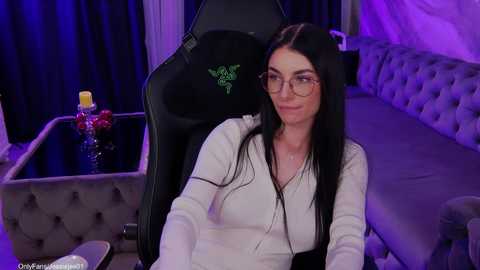A video of a woman with long black hair, glasses, and a white blouse, seated in a black gaming chair, in a dimly lit, purple-lit room with a couch and a table with a vase.
