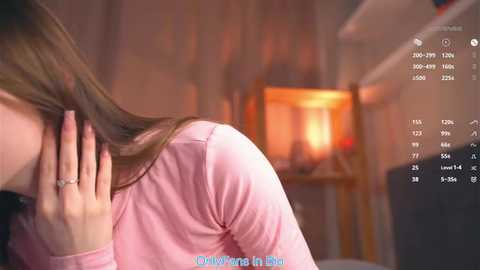 Video of a woman with long brown hair, wearing a pink top, leaning forward with hand on face, in a dimly lit room with blurred, warm-toned background and text overlay.