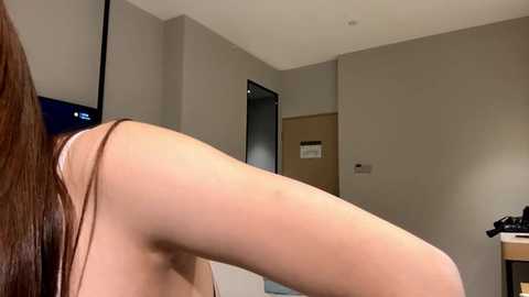 Video of a naked woman with long brown hair bending over, exposing her bare buttocks, in a sparsely furnished beige-walled room with a TV on the left and a door on the right.