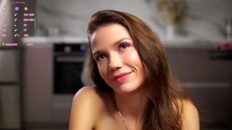 Video of a smiling, topless, brunette woman with long hair, wearing a gold necklace, in a modern kitchen with blurred appliances and a white countertop in the background.