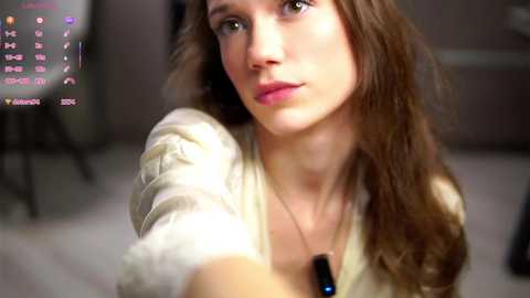 Video of a young Caucasian woman with long brown hair, wearing a cream-colored sweater, holding a black dog tag, in a dimly lit room with a blurred TV screen showing a calendar.