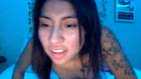 Video of a young, dark-haired woman with tattoos on her arms, wearing a white top, in a dimly lit bedroom with blue lighting.