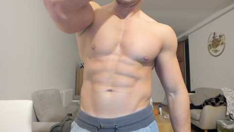 Video of a shirtless, muscular white man with fair skin, showing off his six-pack abs, wearing gray sweatpants, standing in a modern living room with beige furniture.