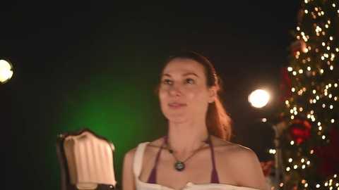 Video of a fair-skinned woman with long, brown hair in a ponytail, wearing a purple dress and necklace, standing in front of a lit Christmas tree.