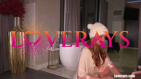 Video of a woman in a cream satin robe, wearing a white knit hat, sitting on a white rug in a modern living room. \"LOVEWAYS\" logo in pink, yellow, and orange text on the background.