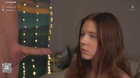 Video of a young woman with long brown hair, looking up at a man's erect penis. Background includes dimly lit bedroom with string lights.