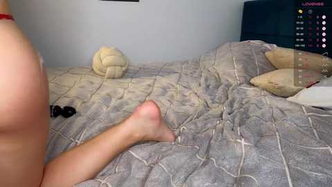 Video of a nude woman lying on a bed with a quilted gray bedspread, a beige ball nearby, and a dark headboard.