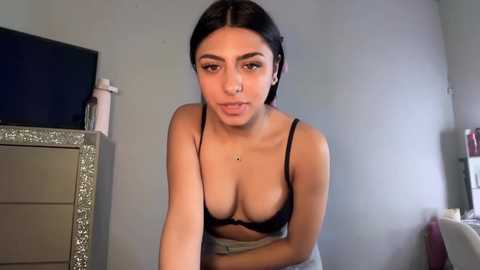 Video of a young Latina woman with dark hair and medium skin tone, wearing a black bra and grey skirt, leaning forward in a dimly lit room with a TV and silver dresser.