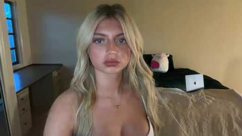 Video of a young woman with long, wavy blonde hair, wearing a white strapless top, standing in a dimly lit bedroom. Background includes a bed with a gray blanket and a laptop, and a window with blue light streaming in.