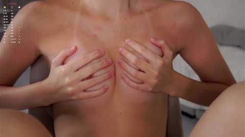 Video of a topless woman with light skin, medium-sized breasts, covering them with her hands, sitting on a bed with a grey pillow in the background.