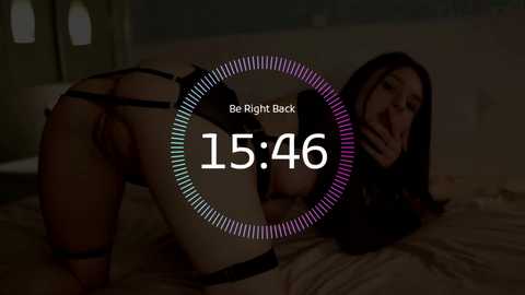 Video of a woman in black lingerie, on her knees on a bed, with a dimly lit room in the background. Text overlay: \"15:46 Be Right Back.\
