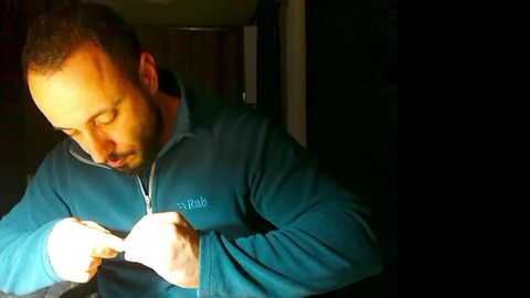 Video of a bearded man in a teal jacket, adjusting his glasses with a gloved hand. Background is dimly lit, with a wooden door partially visible.