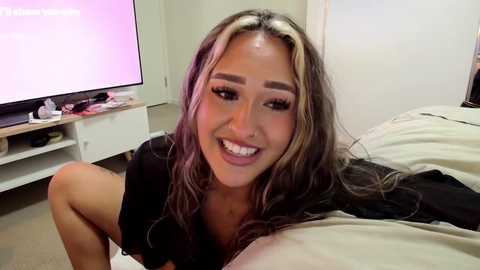Video of a smiling, attractive woman with long, wavy blonde hair, wearing a black top, sitting on a bed in a simple bedroom.