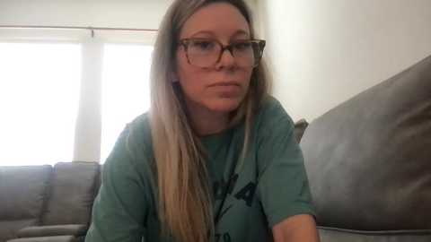 Video of a Caucasian woman with long, straight blonde hair, wearing glasses, a green T-shirt, and sitting on a gray couch in a brightly lit room with white walls and large windows.