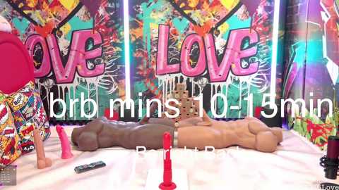 Video of a colorful, graffiti-covered room with two life-size, realistic male sex dolls, one brown and one tan, lying side-by-side on a bed. Various sex toys, including a red dildo and a pink vibrator, are scattered around.