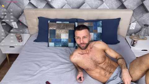 Video of a muscular, bearded, light-skinned man with short brown hair lying shirtless on a bed with grey bedding, blue and beige patterned pillows, and a grey tufted headboard.