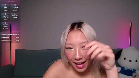 Video of a topless Asian woman with long, platinum blonde hair, wearing pink lipstick, smiling with her finger near her eye, on a gray couch, with a plush toy in the background.
