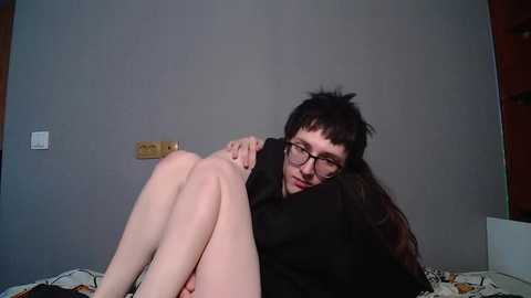 A video of a pale-skinned, long-haired woman with glasses, wearing a black shirt, sitting on a bed with patterned sheets, in a dimly lit room with grey walls.