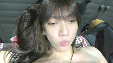 Video of a young Asian woman with long, wavy black hair, pigtails, and a pouty face, wearing a necklace, sitting in a car.