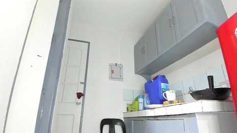 Video of a small, cluttered kitchen with light gray cabinets, white tiled backsplash, a red fridge, and a black stool.