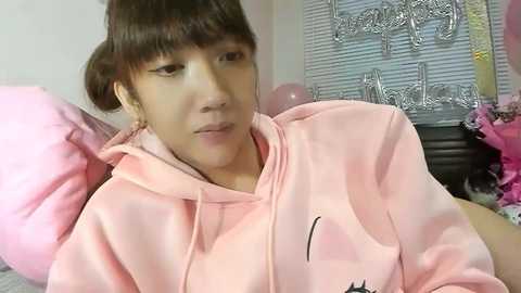 Video of a young Asian woman with straight, dark brown hair in pigtails, wearing a light pink hoodie. She is seated indoors, possibly in a bedroom, with pink bedding and a partially visible window with closed blinds in the background.