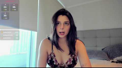 Video of a young woman with long black hair, wearing a black lace bra, sitting on a bed in a modern, minimalist bedroom.