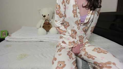 Video of a person in teddy bear pajamas, sitting on a bed with a teddy bear, a green and white pillow, and a white blanket.
