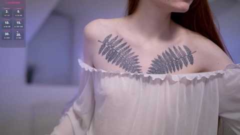 Video of a light-skinned woman with long, straight red hair, wearing a white off-shoulder blouse with a silver fern tattoo on her chest. Background is blurred, featuring a modern, minimalist interior with soft lighting.