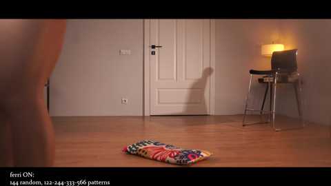 A video of a minimalist bedroom with a wooden floor, featuring a colorful rug, a lit table lamp, and a closed white door.
