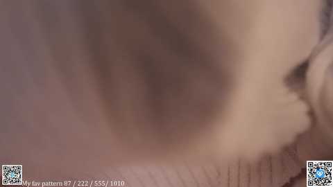 A blurred video of a person's face, possibly a woman, with indistinct features, likely due to motion blur. The background is a neutral, out-of-focus beige. QR codes and text appear at the bottom.
