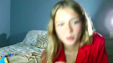 A blurry video of a blonde woman with light skin, wearing a red shirt, sitting on a bed with a beige blanket and patterned pillows in a dimly lit room.