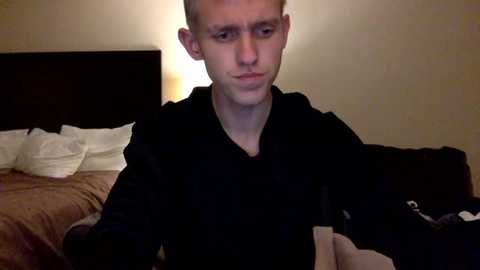 Video of a young, fair-skinned man with short, light brown hair, wearing a black long-sleeve shirt, sitting on a bed with beige sheets and a dark headboard, holding a small, light-colored object.