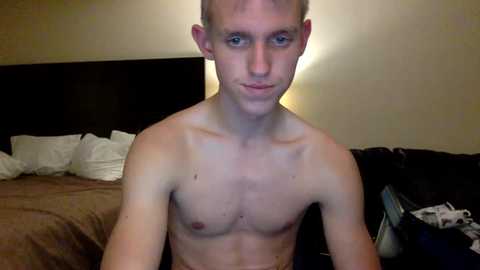 Video of a shirtless, lean, young Caucasian man with short blond hair, blue eyes, and fair skin, sitting on a bed with a brown comforter. The background features a dark headboard, white pillows, and a lamp.