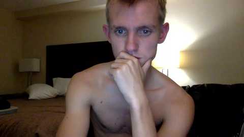 Video of a shirtless, fair-skinned, slender young man with short brown hair, blue eyes, and a contemplative expression, sitting on a bed in a dimly lit, beige-walled room with a bedside lamp.