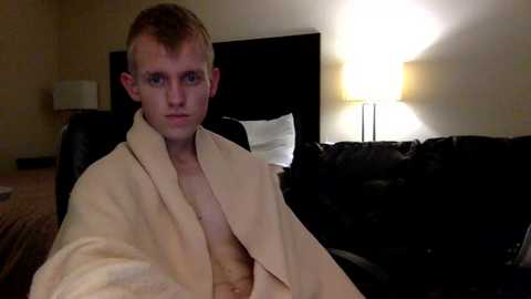 Video of a young, fair-skinned, short-haired man in a beige bathrobe, sitting on a black leather couch in a dimly-lit room with a white lamp and beige walls.